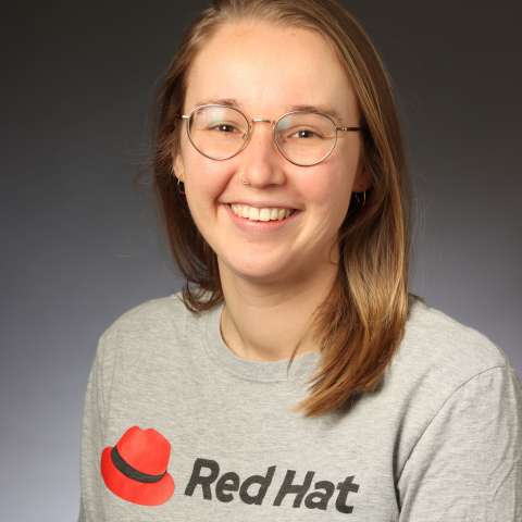 A professional headshot of Kathryn who has light brown hair and wire frame glasses wearing a Red Hat shirt